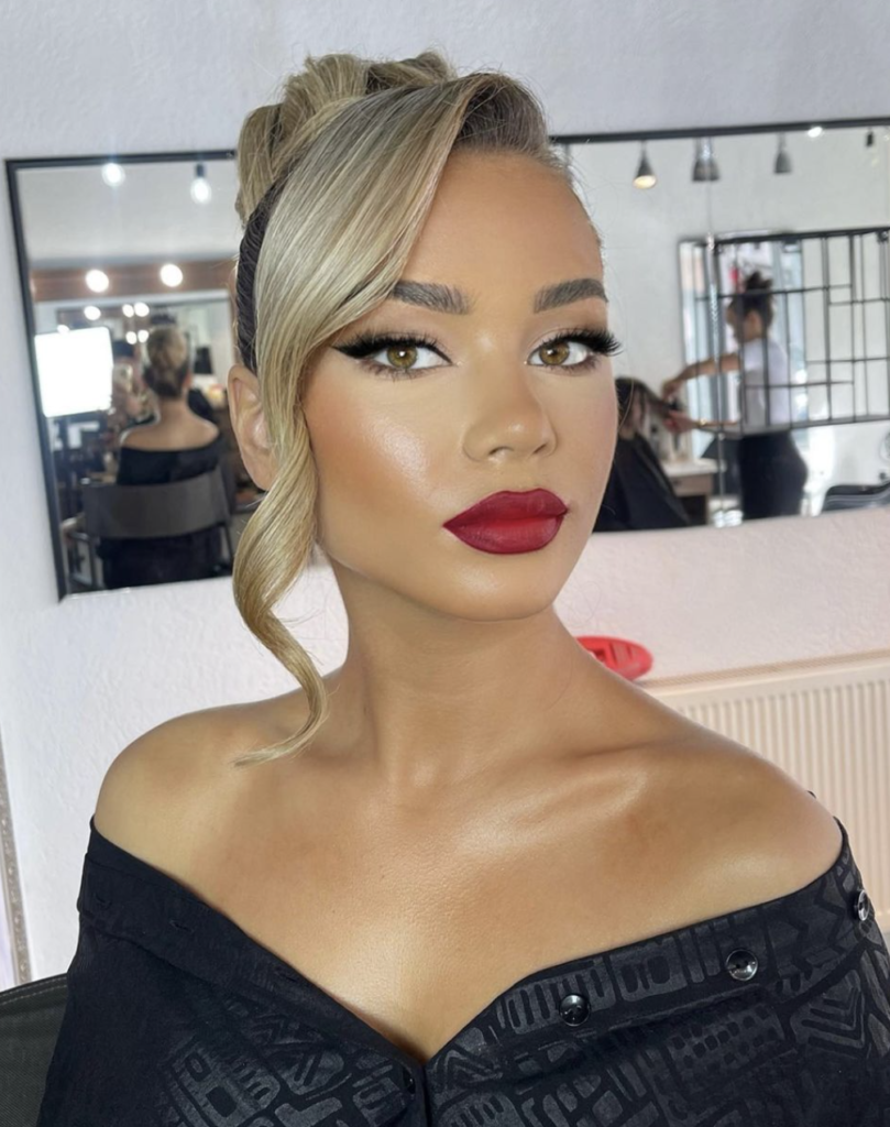 This image features a woman sporting a bold, classic red lip makeup look. Her eyes are defined with winged eyeliner and fluttery lashes, while her brows are perfectly sculpted. She exhibits a glowing complexion with a touch of bronzer and highlighter. Her blonde hair is styled into an elegant updo, and she wears a black off-the-shoulder top.